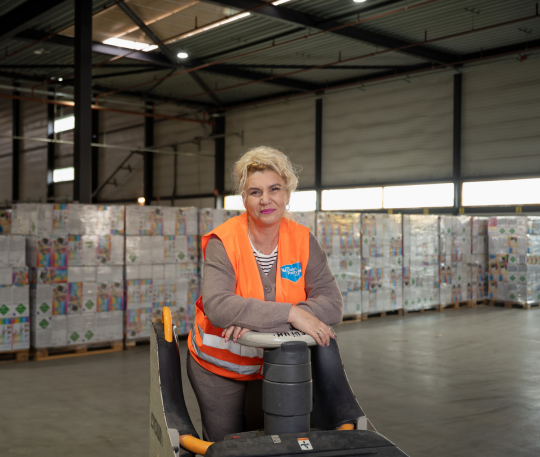 Magazijnmedewerker staat bij een elektrische pallettruck met gestapelde goederen op de achtergrond.